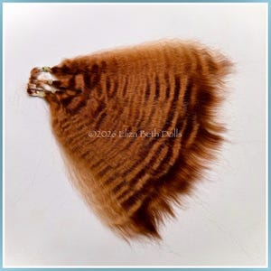 May include: A fan-shaped arrangement of ginger-colored doll hair wefts featuring a crimped texture and dark horizontal stripes. The hair is secured at the base with small bands. The text 2026 Eliza Beth Dolls is visible in the center.