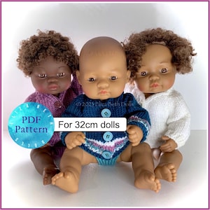 May include: Three knitted doll sweaters in pink, blue, and white for 32 cm dolls. The sweaters are shown on three dolls with different skin tones.