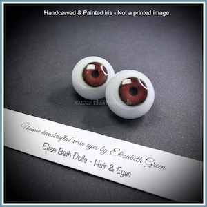 May include: Two spherical resin doll eyes featuring hand-carved and painted reddish-brown irises with black pupils. The text reads: Handcarved & Painted iris - Not a printed image, ©2026 Eliza Beth Dolls, Unique handcrafted resin eyes by Elizabeth Green.