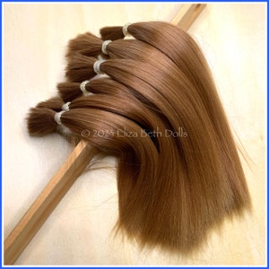 May include: A collection of light brown doll hair extensions, each bundled with a clear elastic band. The hair is arranged on a wooden support, showcasing its smooth texture and color. The image is set against a light-colored background.