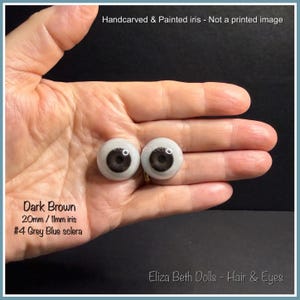 May include: Two round doll eyes with hand-carved and painted dark brown irises and grey-blue sclera. The text reads: Handcarved & Painted iris - Not a printed image, Dark Brown, 20mm / 11mm iris, #4 Grey Blue sclera, Eliza Beth Dolls - Hair & Eyes.