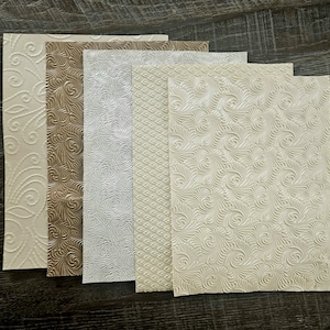 May include: A selection of textured paper sheets in neutral tones. The papers display embossed patterns, such as swirls, floral designs, and geometric shapes. The colours vary from cream and beige to light brown and white.