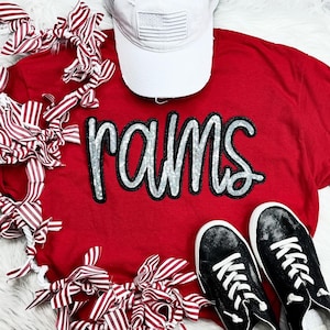 May include: Red t-shirt with the word "rams" in silver glitter letters. The shirt is laying on a white surface with red and white striped ribbon.
