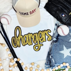 May include: White t-shirt with the word "Chargers" in gold glitter letters. The shirt is paired with a beige baseball cap that says "Game Day" in maroon letters. The image also includes a pair of denim shorts with a white star design and baseballs.