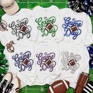 May include: Six white long-sleeve sweatshirts featuring a sequined graphic of a football and the text Game Day in various colours including black, green, blue, navy, purple, and silver, arranged on a green field-patterned surface.