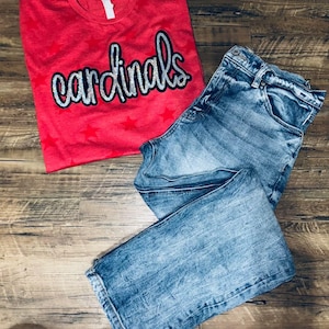 Può includere: Una maglietta rossa con la parola "cardinals" in lettere argentate glitter. La maglietta ha delle stelle bianche stampate. Un paio di jeans blu sono sopra la maglietta.