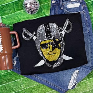 Op de afbeelding: Zwart T-shirt met een pailletten Raiders-logo met helm, zonnebril en gekruiste zwaarden. Een bruine beker met voetbalthema en een discobal zijn ook zichtbaar.