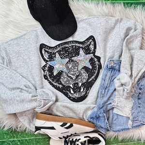 May include: A light gray sweatshirt with a black and silver glitter panther graphic wearing star-shaped sunglasses. The image also includes a black corduroy baseball cap, denim shorts, and white sneakers with brown soles.