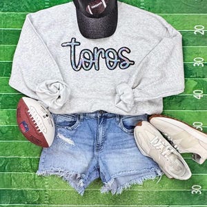 May include: A light gray sweatshirt with the word "toros" in a glittery font, paired with denim shorts, sneakers, and a black baseball cap with a football design. A football is also included.