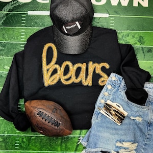 Peut inclure: Sweat-shirt noir avec inscription "Bears" en paillettes dorées, casquette de camionneur noire en mesh avec un motif de ballon de football, ballon de football en cuir marron et short en jean avec des bracelets dorés et noirs.