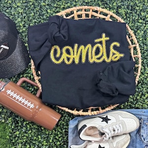 Puede incluir: Una sudadera negra con la palabra "comets" en letras doradas con purpurina se muestra en una cesta tejida. También se ven una gorra de béisbol negra, un vaso marrón con forma de balón de fútbol y zapatillas blancas con estrellas negras.