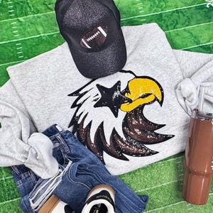 Könnte beinhalten: Ein graues Sweatshirt mit einem paillettenbesetzten Adlerdesign, eine schwarze Baseballkappe mit einem Football, blaue Jeans und ein brauner Becher. Das Sweatshirt zeigt einen großen Adlerkopf mit gelben, braunen und weißen Pailletten.
