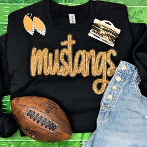 Könnte beinhalten: Schwarzes Sweatshirt mit dem Wort "mustangs" in goldenen Pailletten. Das Bild zeigt einen Football, Jeansshorts, Ohrringe und Armbänder. Das Sweatshirt ist von der Marke Gildan.