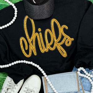 May include: Black long-sleeve sweatshirt with a gold glitter "Chiefs" design. The sweatshirt is paired with a black baseball cap and a white pearl necklace.