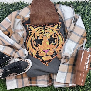 Könnte beinhalten: Ein lässiges Outfit mit einem dunkelgrauen T-Shirt mit einem paillettenbesetzten Tigergesicht, einer braunen Mütze und einem karierten Flanellhemd. Schwarze Sneaker mit Leopardenmuster und ein brauner Becher vervollständigen das Ensemble.