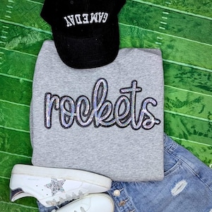 Può includere: Una felpa grigia con la parola "rockets" in lettere nere e argento glitter. La felpa è abbinata a jeans blu e scarpe da ginnastica bianche con una stella argentata.