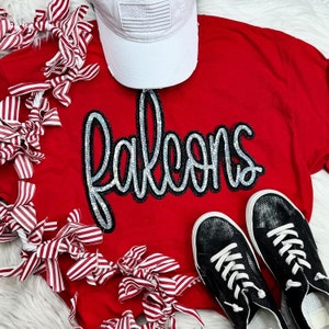 Könnte beinhalten: Rotes T-Shirt mit einer weißen Baseballkappe und einem glitzernden schwarzen und silbernen "Falcons"-Design. Das Shirt ist von einem rot-weiß gestreiften Band umgeben.