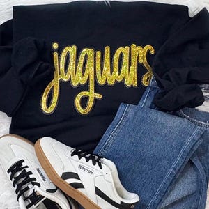 May include: Black sweatshirt with the word "jaguars" in gold glitter lettering. The sweatshirt is paired with blue jeans and white Reebok sneakers with black accents and brown soles.