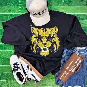 May include: A black sweatshirt with a gold sequined tiger design, paired with a "Game Day" baseball cap, white sneakers, a football-themed tumbler, and blue jeans. The items are arranged on a green surface.