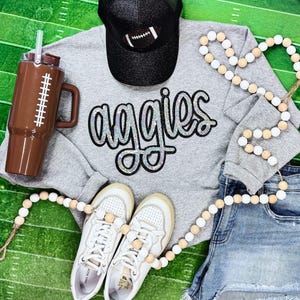 Puede incluir: Una sudadera gris con la palabra "Aggies" en letras plateadas brillantes. La sudadera se combina con zapatillas blancas, pantalones cortos de mezclilla, una gorra de béisbol negra con brillantina y un collar de cuentas blancas y marrones.