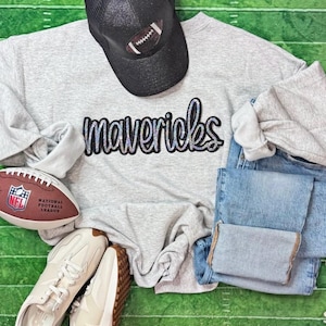 May include: A grey sweatshirt with the word "Mavericks" in a sparkly font, a black baseball cap with a football design, a football, jeans, and trainers are arranged on a green field background.