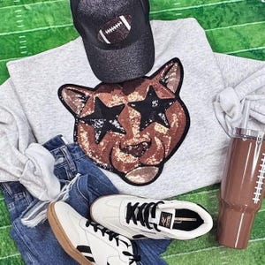 May include: A casual outfit featuring a gray sweatshirt with a sequined cat face design, a black baseball cap with a football, blue denim shorts, white sneakers, and a brown tumbler with a football design. The items are arranged on a green field.