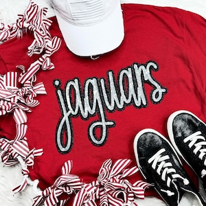 May include: Red t-shirt with the word "Jaguars" in black and silver glitter lettering. The shirt is surrounded by red and white striped fabric.