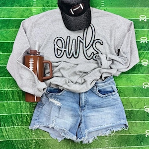 May include: A gray long-sleeve sweatshirt with the word "owls" in a script font, paired with denim shorts and a black baseball cap featuring a football design. A brown tumbler with a football graphic is also included.