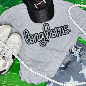 May include: A gray t-shirt with a black and silver glitter "Longhorns" design. The shirt is paired with a black baseball cap and a white beaded necklace.