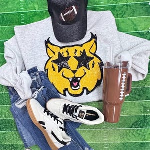 May include: A gray sweatshirt with a yellow cat graphic, a black baseball cap with a football, blue jeans, white sneakers, and a brown football-themed tumbler with a straw. The items are arranged on a green field.