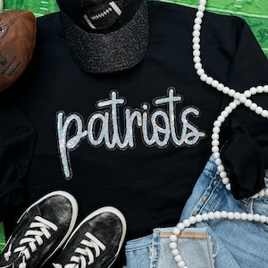 Könnte beinhalten: Schwarzes Sweatshirt mit einem silbernen Pailletten-Aufdruck "patriots". Das Sweatshirt wird mit einer schwarzen Baseballkappe, einer weißen Perlenkette und einer blauen Jeans kombiniert.
