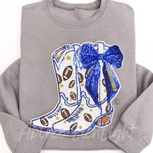 Op de afbeelding: Een grijze sweatshirt met een groot, versierd ontwerp van witte cowboylaarzen versierd met voetballen en gouden sterren. Een blauwe strik met gouden accenten is aan de laarzen bevestigd. Het ontwerp is gemaakt met pailletten en kralen.