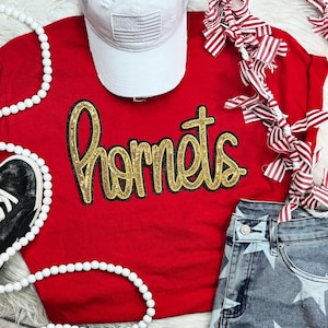 May include: Red t-shirt with a gold glitter "Hornets" design. The shirt is paired with a white baseball cap, a white beaded necklace, and blue denim shorts with white stars.