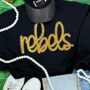 May include: Black sweatshirt with a gold glitter "rebels" applique. The sweatshirt is laid flat on a green and white surface with a black baseball cap, white pearl necklace, and a pair of white sneakers.