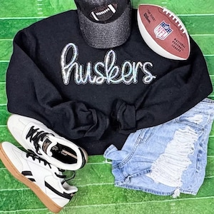 Könnte beinhalten: Ein schwarzes Sweatshirt mit dem Wort "huskers" in schillernder Schrift, eine graue Baseballkappe mit Football-Design, ein Football, zerrissene Jeansshorts und weiße Turnschuhe mit schwarzen Akzenten und braunen Sohlen, angeordnet auf einer grünen Oberfläche.
