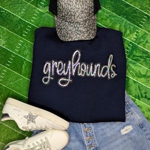 May include: A navy blue sweatshirt with the word "Greyhounds" in silver glitter lettering. The sweatshirt is paired with a light blue denim skirt and white sneakers.