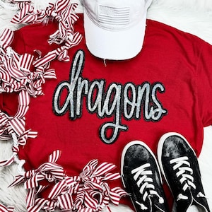 Könnte beinhalten: Rotes T-Shirt mit einem schwarzen und silbernen Glitzer-Bügelbild, auf dem "dragons" steht. Das Shirt liegt auf einer weißen Oberfläche mit rot-weiß gestreiftem Stoff.