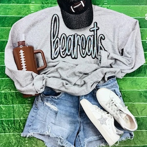 May include: A gray sweatshirt with the word "bearcats" in sparkly silver letters. The sweatshirt is worn with denim shorts and white sneakers with silver glitter.