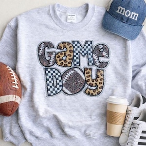 May include: A light gray sweatshirt with the words "Game Day" in a football-themed design. The letters have a mix of football, leopard print, and checkered patterns. A denim baseball cap with "mom" embroidered on it is also visible.