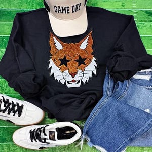 May include: Black sweatshirt with a sequined bobcat face design, a beige "GAME DAY" baseball cap, blue denim jeans, and white sneakers with black accents. The bobcat has star-shaped eyes. The items are arranged on a green surface.