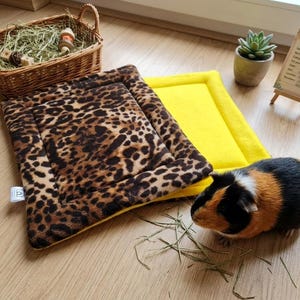 May include: A rectangular pet mat featuring a brown and black leopard print on one side and a solid bright yellow color on the other. A tricolor guinea pig with black, brown, and white fur stands next to the mat, near a wicker basket of hay.