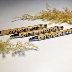 Könnte beinhalten: Drei Holzstift mit silbernen Akzenten und schwarzer Tinte. Die Stifte haben folgenden Text: "Raise Your Horns", "Drenngr of Ragnarok" und "Victory or Valhalla".