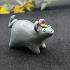 May include: A gray ceramic mouse figurine with pink accents on its ears and feet. The mouse has a small green and yellow decoration on its head. The figurine is on a dark gray surface.