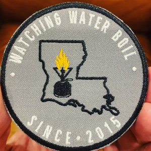 May include: A gray embroidered patch with a black outline of the state of Louisiana. The patch has the text "Watching Water Boil Since 2015" around the outside and a black silhouette of a pot with a yellow flame inside.