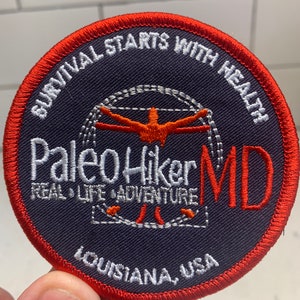May include: A round, red-bordered patch with a dark blue background. The patch features white text that reads "Survival Starts With Health" and "Paleo Hiker MD Real Life Adventure" in a white and red font. The text "Louisiana, USA" is also visible in white text at the bottom of the patch.