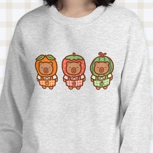 K&ouml;nnte beinhalten: Ein graues Sweatshirt mit einem Aufdruck von drei braunen B&auml;ren, die Fruchtkost&uuml;me tragen. Die B&auml;ren tragen ein orangefarbenes, rotes und gr&uuml;nes Fruchtkost&uuml;m. Das Sweatshirt hat einen Rundhalsausschnitt und lange &Auml;rmel.