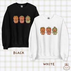 K&ouml;nnte beinhalten: Zwei Sweatshirts, eines schwarz und eines wei&szlig;, mit einem Motiv von drei Cartoon-B&auml;ren, die Fruchtkost&uuml;me tragen. Die B&auml;ren tragen orange, rote und gr&uuml;ne Fruchtkost&uuml;me. Der Text "BLACK" und "WHITE" befindet sich unter jedem Sweatshirt.