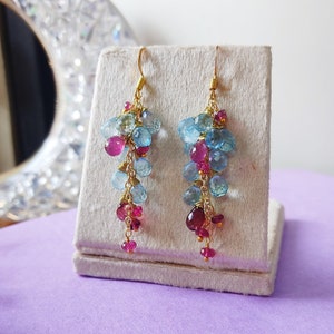 Peut inclure: Une paire de boucles d'oreilles pendantes avec une chaîne dorée et des pierres précieuses bleues et roses. Les boucles d'oreilles sont présentées sur un support en velours blanc.