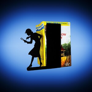 May include: A black metal bookend featuring a silhouette of a woman holding a magnifying glass, positioned as if investigating. The bookend is placed between a stack of yellow and white books, with the title "Nancy Drew" visible.