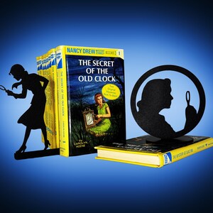 May include: A display featuring vintage Nancy Drew mystery books, including "The Secret of the Old Clock." Black silhouette bookends depict a woman with a magnifying glass. The books have yellow spines and colorful covers.